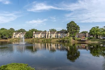 A serene lake with a fountain in the middle, surrounded by houses and trees under a clear blue sky.